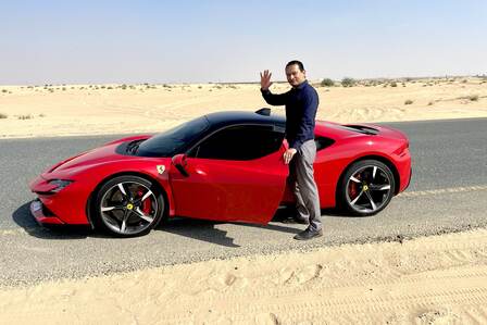 Gustavo saludando junto a un Ferrari rojo, en una carretera del desierto de Sharjah